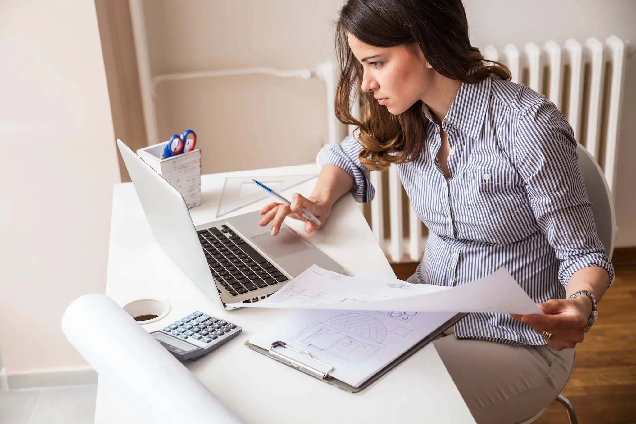 A focused woman working on a laptop at a white desk with papers, a calculator, and office supplies.