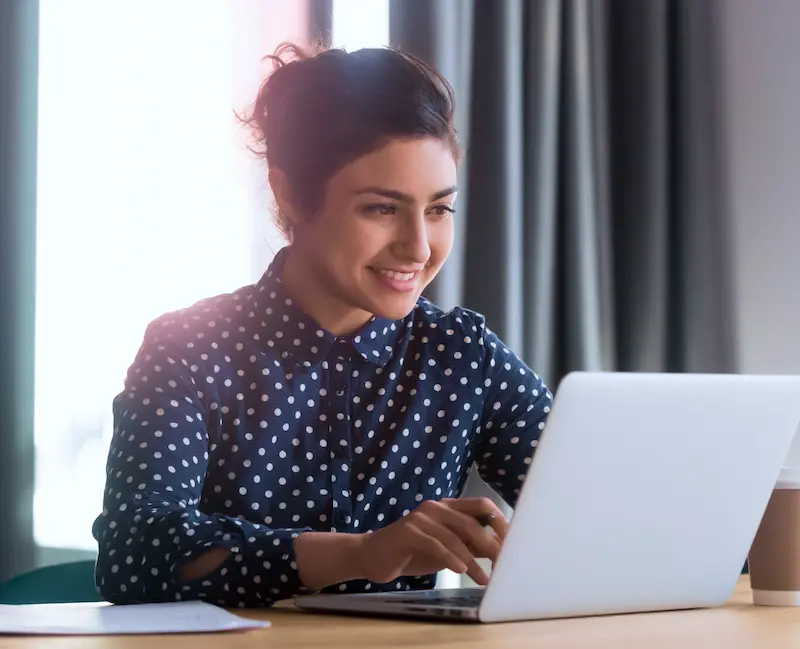 Woman in a polka dot shirt working on a laptop at a desk, beside a cup of coffee.