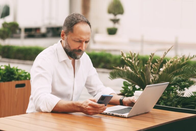 Man working outdoors on a laptop and smartphone surrounded by plants.