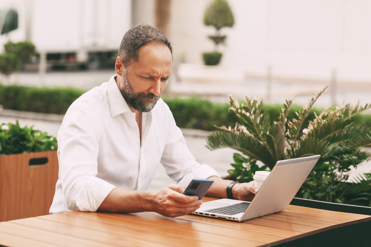 Man working outdoors on a laptop and smartphone surrounded by plants.