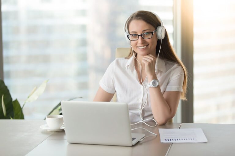 A smiling woman uses a laptop with headphones in a bright office setting, indicating effective virtual assistant work.