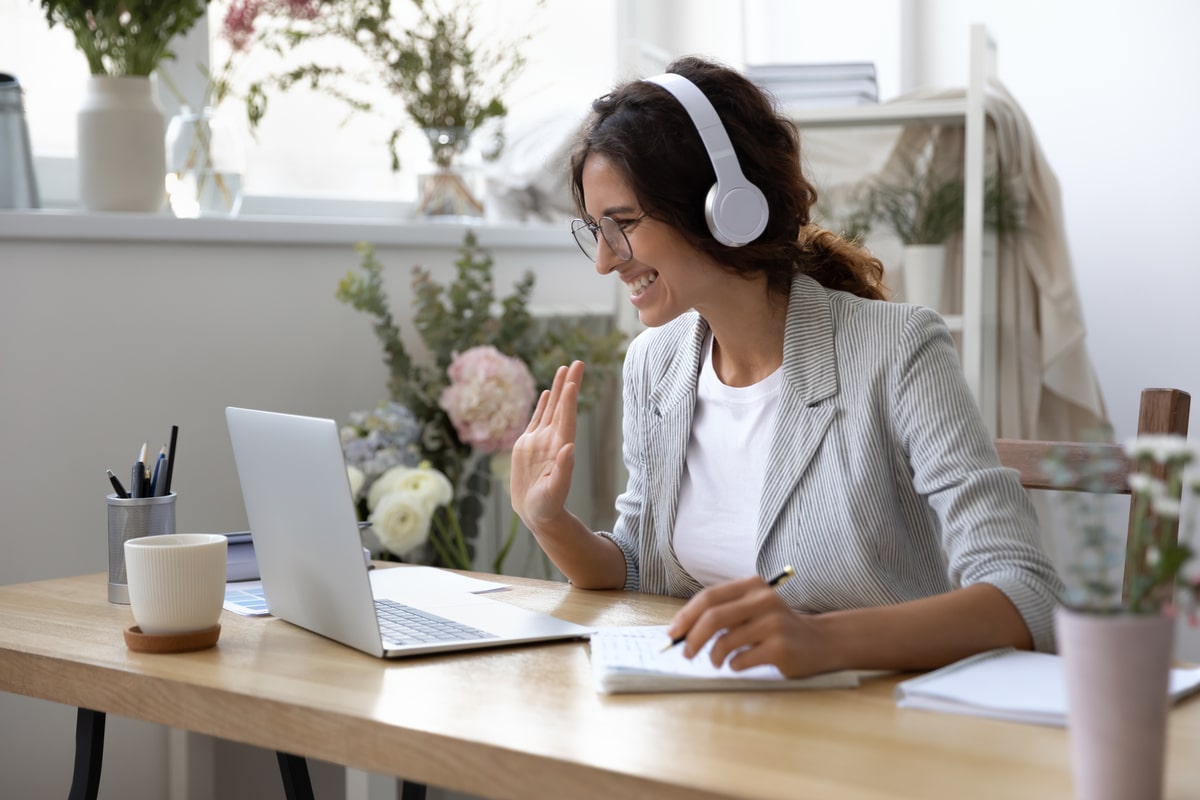 Woman wearing headphones smiling at laptop while working from home office desk, surrounded by plants.