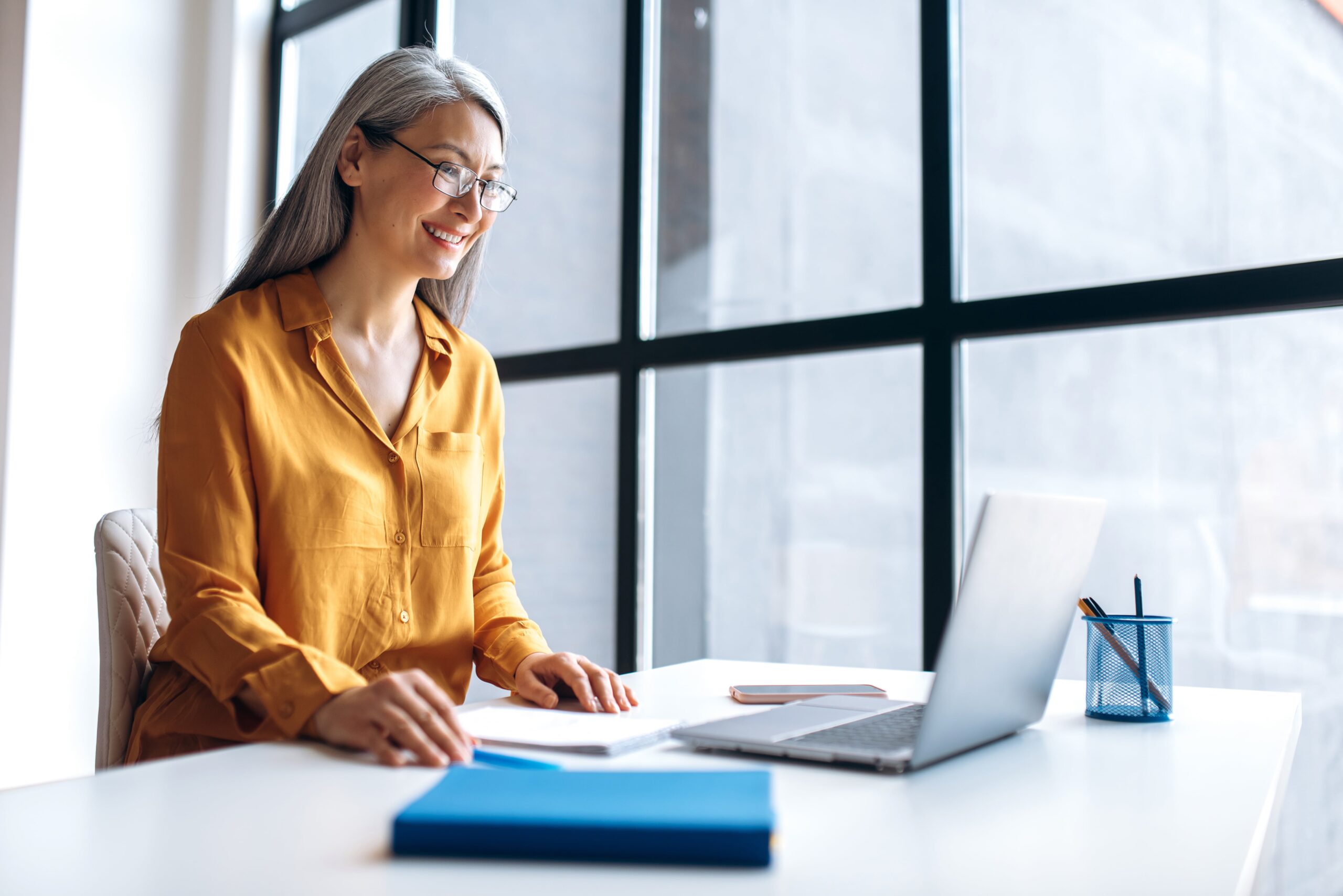 A smiling professional woman working on a laptop at a desk in a bright, modern office setting.
