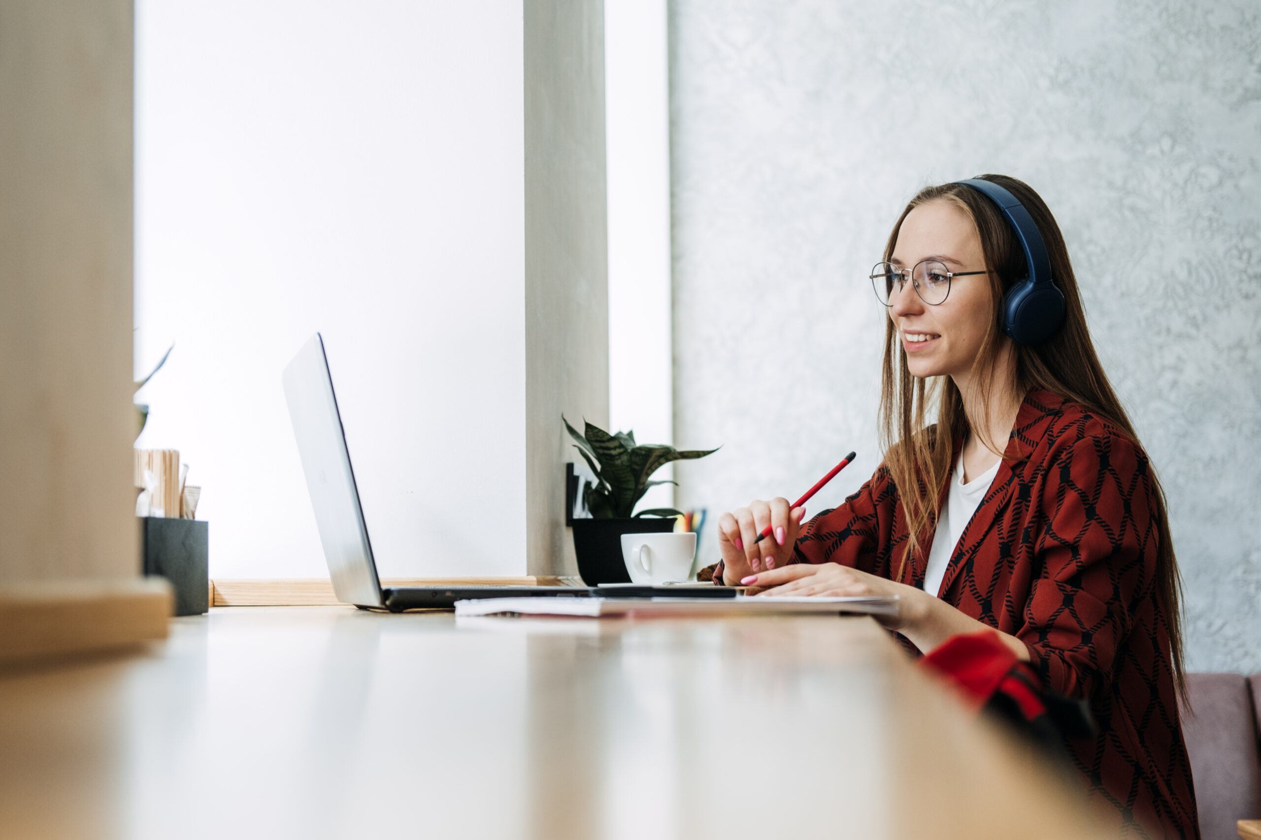 A young woman wearing headphones, working on a laptop and taking notes in a modern workspace.