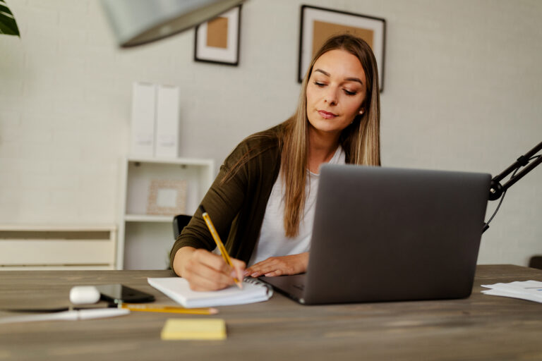 Focused administrative assistant taking notes while working on a laptop in a modern office.