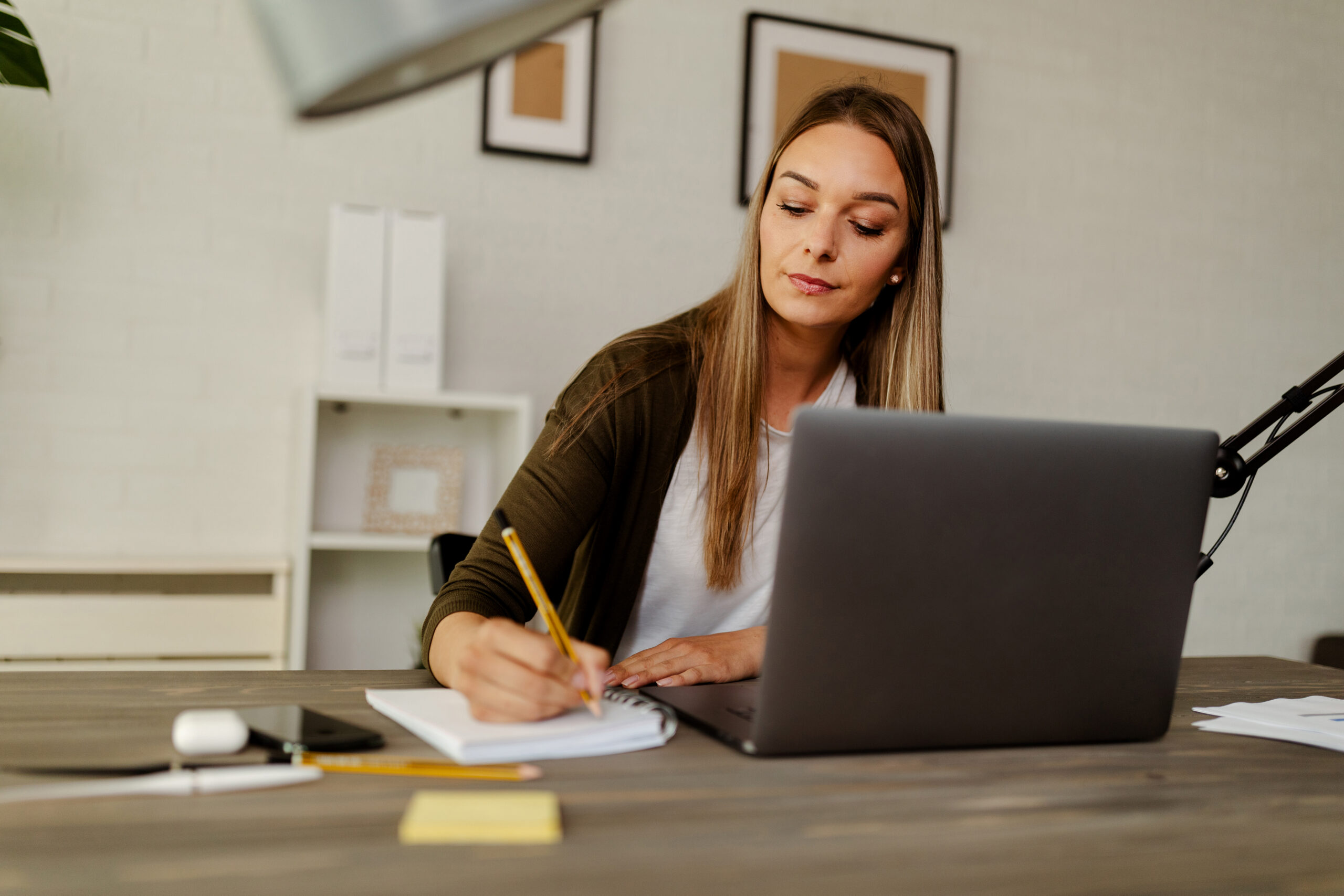 Focused administrative assistant taking notes while working on a laptop in a modern office.