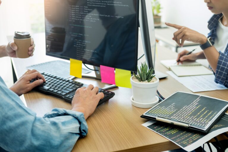 Developers collaborating on code with a computer monitor, notebook, and coffee in a modern workspace.
