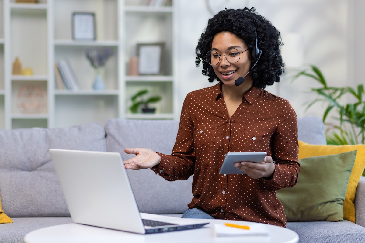African American female teacher with curly hair using a laptop and headset for online in a cozy home setting.