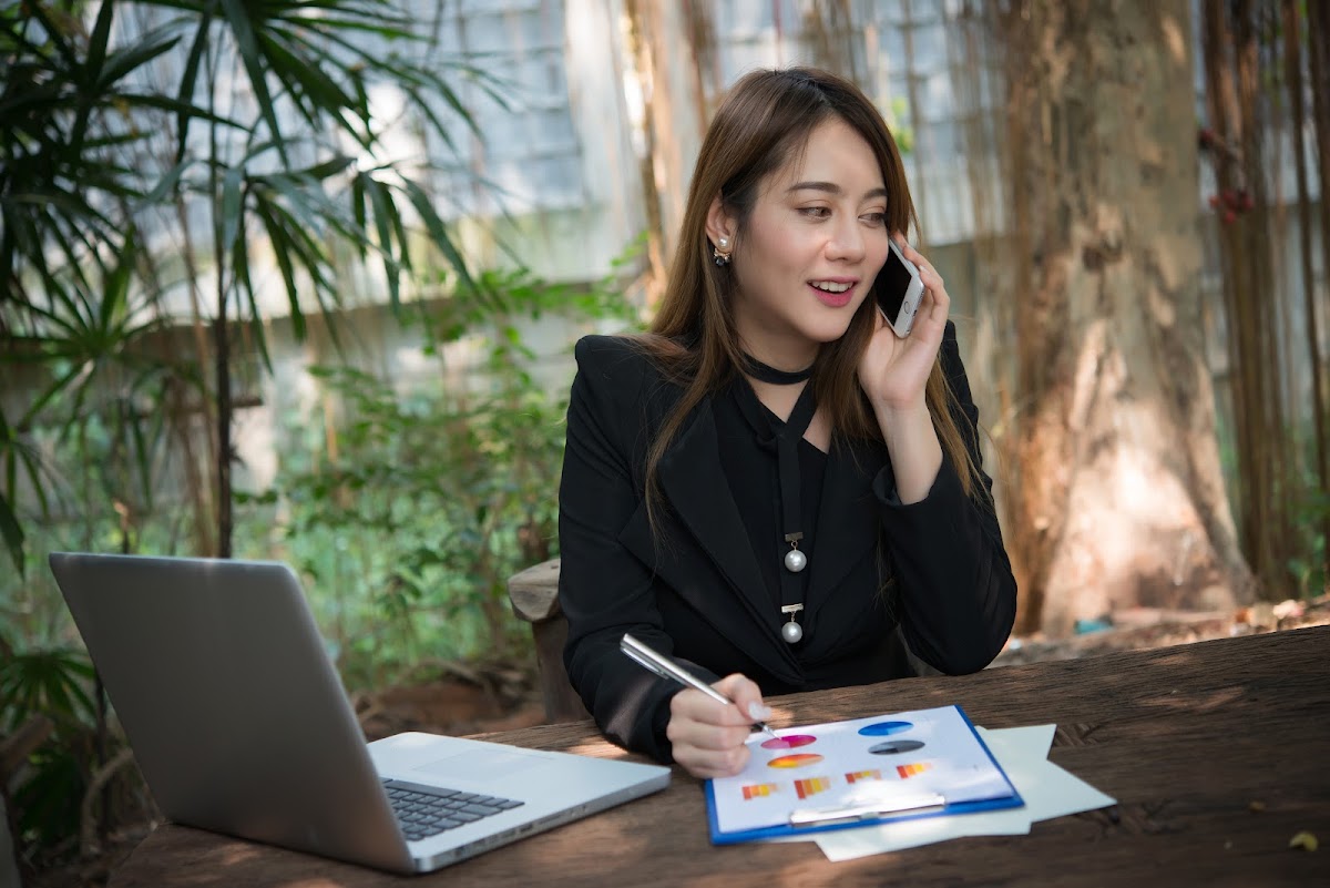 A businesswoman in a black suit talking on the phone while working on a laptop and analyzing data outdoors.