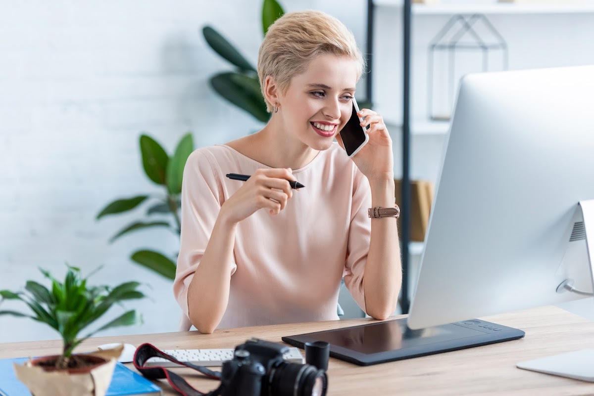 A smiling woman sitting at a desk, holding a pen and talking on the phone while looking at a Computer screen.