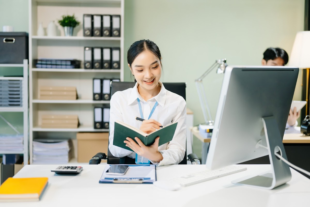 Asian female office worker in a business suit smiling while writing at her desk.