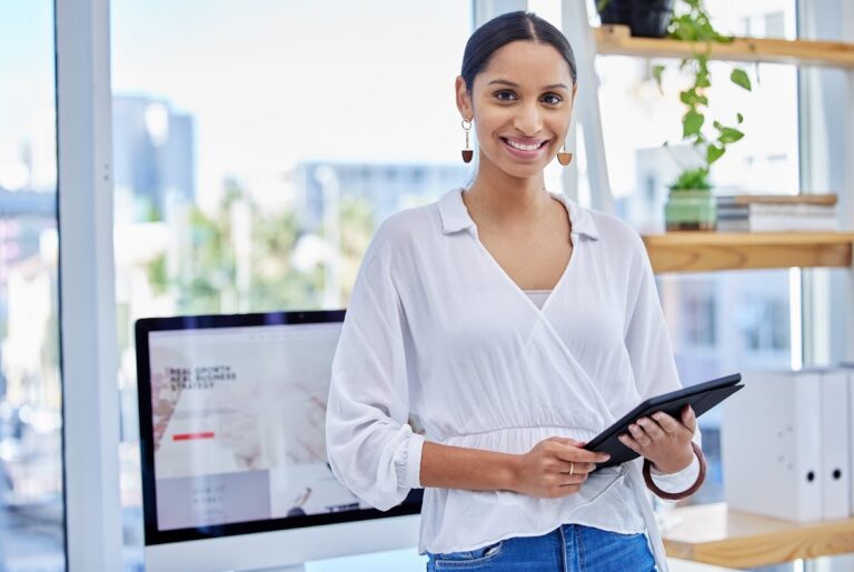 A woman smiling while holding a tablet, standing in a modern office with a computer in the background, demonstrating virtual marketing assistant services.