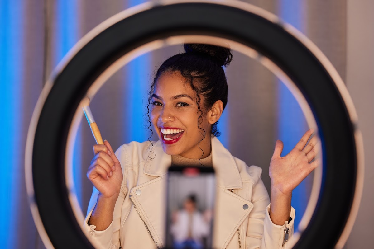 A cheerful woman framed within a ring light, holding a makeup brush and engaging with the camera in a lively content creation setup with blue ambient lighting.