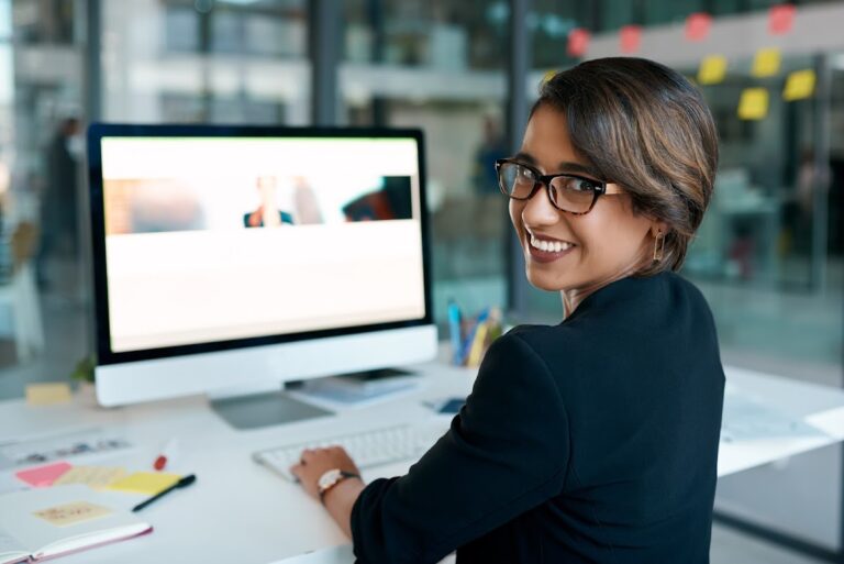 A smiling woman sitting at a desk with a computer, looking back at the camera in a professional office setting.