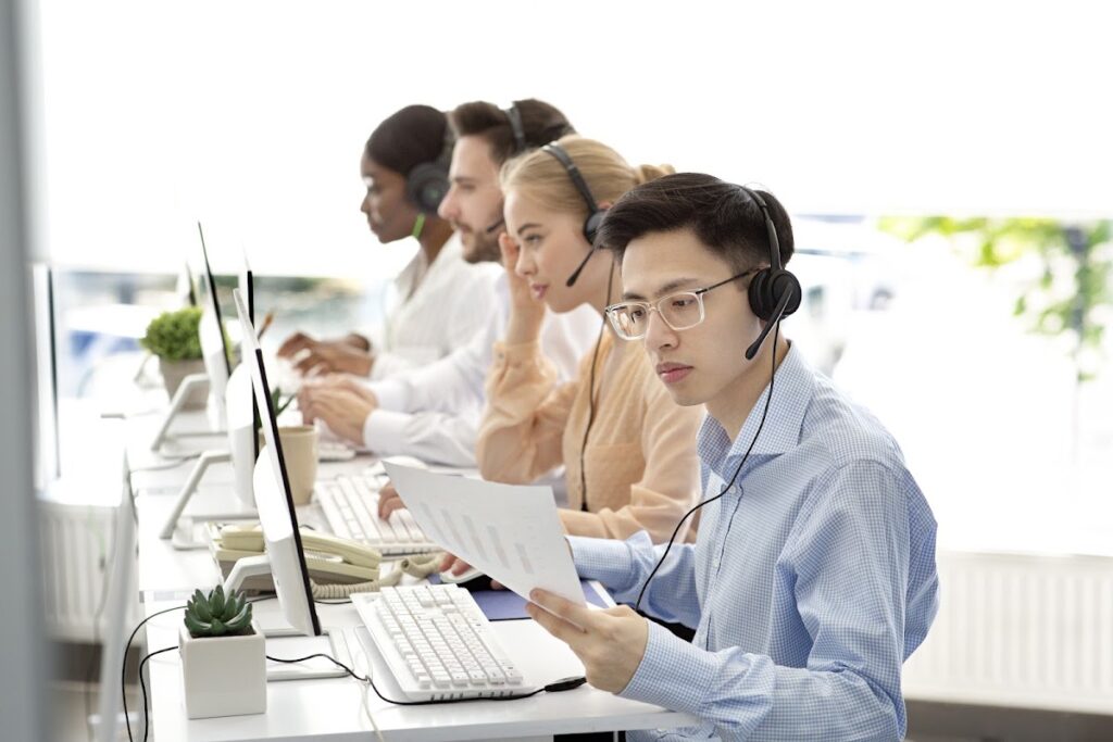 A group of diverse call center agents wearing headsets and working on computers in a well-lit, organized office.
