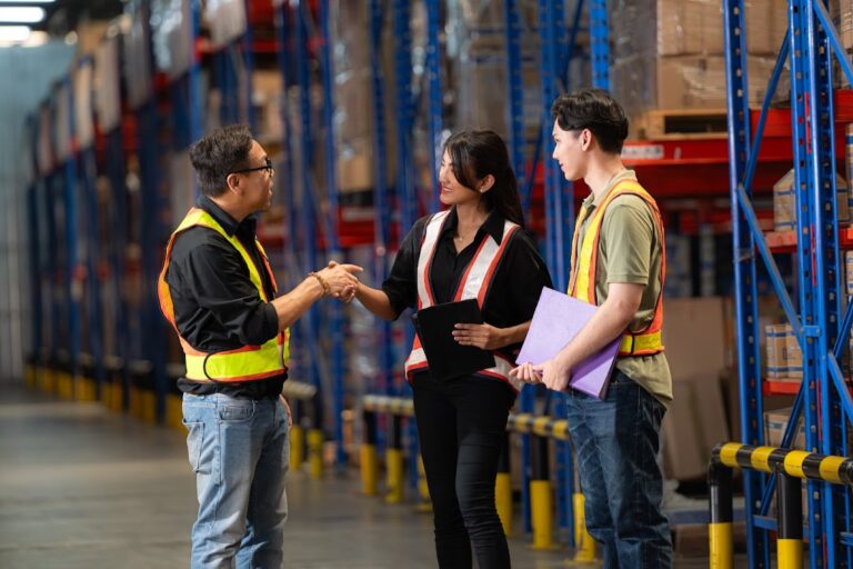 Warehouse team in safety vests discussing product inventory in storage aisle.