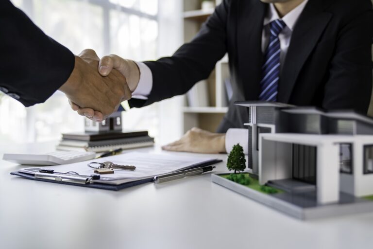 Real estate agent and clients shaking hands across a contract, surrounded by model houses and keys.