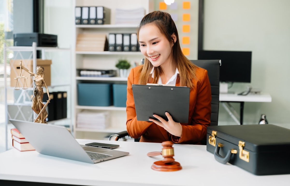 Smiling professional woman in an office with a laptop, clipboard, and legal scales in the background.