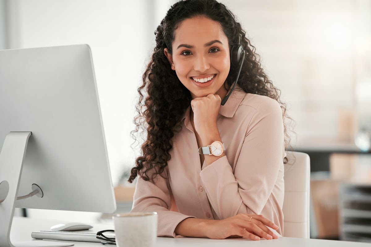 Smiling woman with curly hair wearing a headset, sitting at a desk with a computer, exuding professionalism.