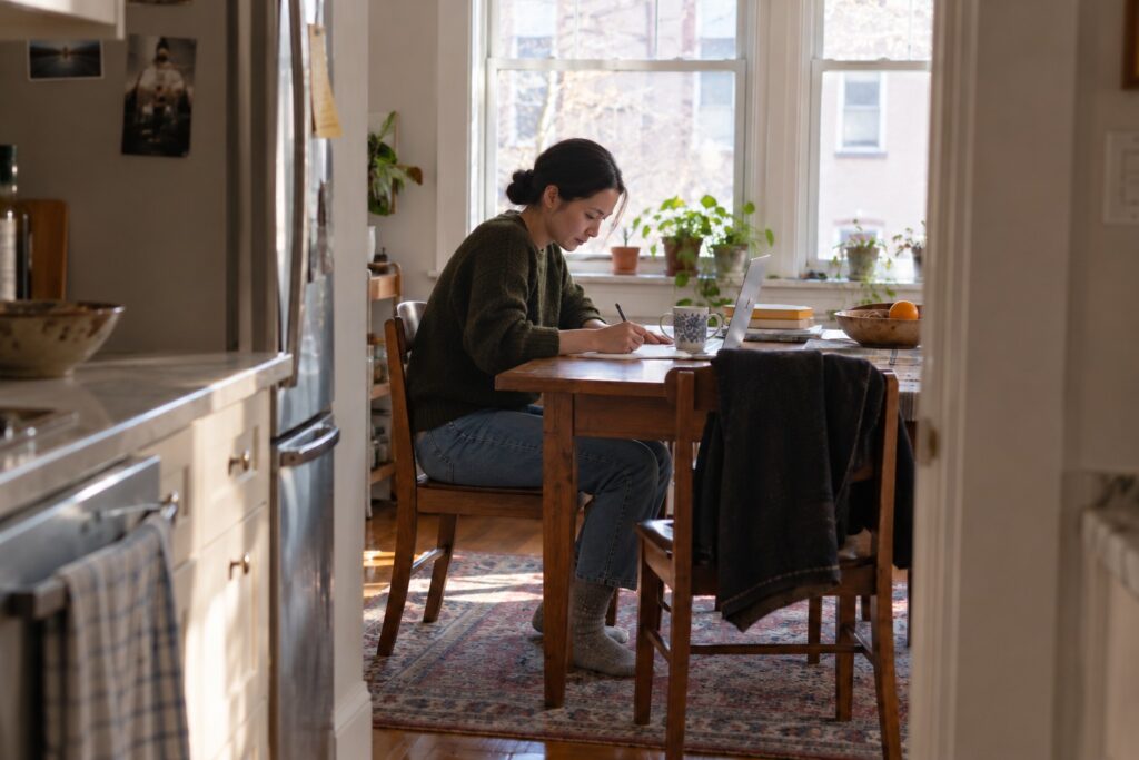 Woman working on a laptop and writing notes at her kitchen table.